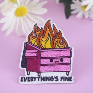 May include: Embroidered patch showing a pink dumpster engulfed in flames, with orange and yellow hues. The text "EVERYTHING'S FINE" is displayed. Daisies are visible in the background.