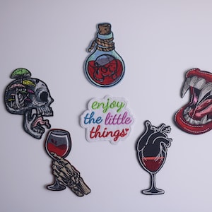 May include: Five embroidered iron-on patches with various designs. One patch features a skull with mushrooms inside, another a bottle with a skull inside, another a heart with a glass of wine, another a mouth with fangs, and the last one says "enjoy the little things" in rainbow colors.