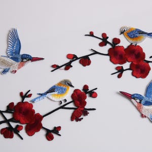 Peut inclure: Écussons brodés représentant des oiseaux colorés et des fleurs rouges sur des branches noires. Les oiseaux ont des plumes bleues, jaunes et blanches avec des becs rouges. Idéal pour les vêtements ou les projets d'artisanat.