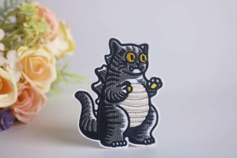 Peut inclure: Patch brod&eacute; thermocollant repr&eacute;sentant un chat noir et blanc aux yeux jaunes, portant une chemise ray&eacute;e et se tenant dans une pose de Godzilla.