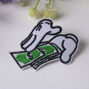 Puede incluir: Parche bordado con manos de dibujos animados blancas sosteniendo dinero verde con contornos negros. El parche está sobre un fondo blanco, con flores moradas y blancas en el fondo.