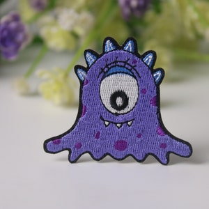 May include: Purple embroidered iron-on patch of a cartoon monster with one large eye, a toothy grin, and purple spots.