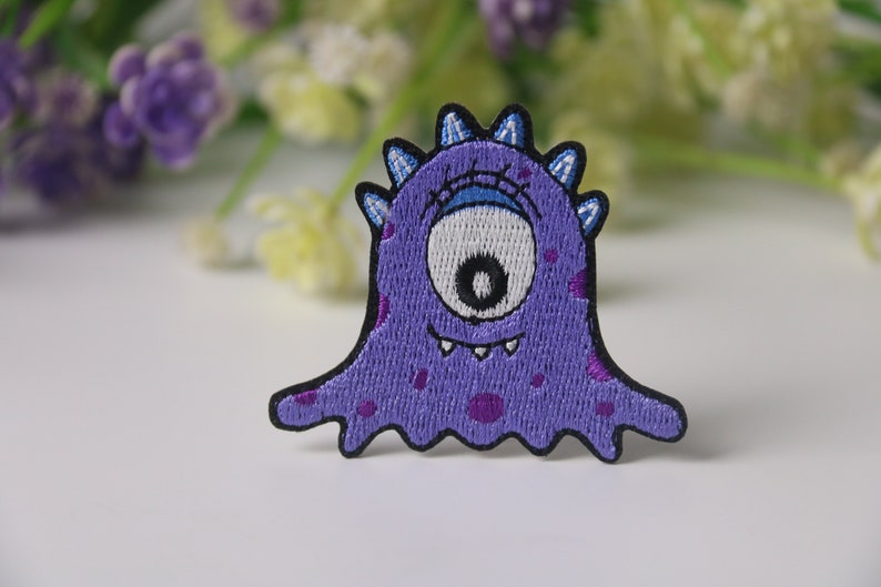 May include: Purple embroidered iron-on patch of a cartoon monster with one large eye, a toothy grin, and purple spots.