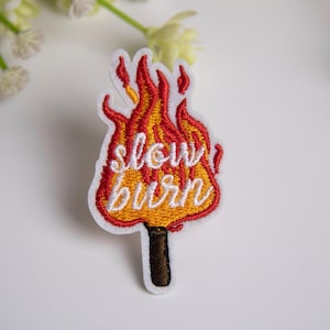 May include: Embroidered patch with a flame design in red, orange, and yellow, and the words "slow burn" in white script. The patch has a white border and a brown stick. The patch is on a white surface.