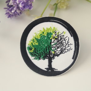 May include: A round black and white embroidered patch with a green and black tree design. The tree has a black trunk and branches with green and yellow leaves.