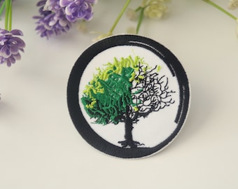 May include: A round black and white embroidered patch with a green and black tree design. The tree has a black trunk and branches with green and yellow leaves.