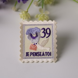 May include: A white embroidered patch shaped like a postage stamp with a purple flower, a white dove carrying a pink envelope, and the text "JE PENSAIS A TOI" and "39".