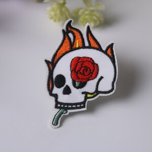 May include: White embroidered patch of a skull with a red rose in its eye socket, set aflame with orange and yellow flames.