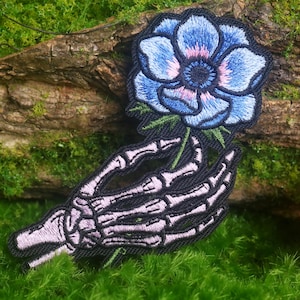 May include: An embroidered patch featuring a light purple skeleton hand holding a blue flower with a dark center. The design is outlined in black thread, showcasing detailed bone structures and delicate blue petals with subtle pink highlights.