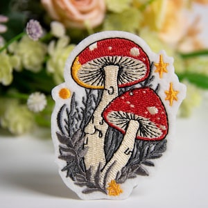 Op de afbeelding: Geborduurde patch met twee rode paddenstoelen met witte stippen en crèmekleurige stelen. De paddenstoelenkappen hebben een gele maandetail. De patch bevat grijs blad en gouden steraccenten, perfect voor jassen, tassen of andere stoffen items.