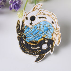 May include: Embroidered patch featuring two koi fish, one black and one white, swimming in a circle. The fish are outlined in gold thread and the background is a blue and white swirl pattern.