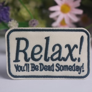 May include: A rectangular embroidered patch with a cream-colored background and a dark navy blue border. The patch features the dark navy blue embroidered text "Relax!" on the top line and "You'll Be Dead Someday!" on the bottom line.
