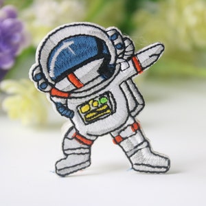Könnte beinhalten: Weißer gestickter Bügel-Patch mit einem Astronauten, der den Dab macht. Der Astronaut trägt einen weißen Raumanzug mit einem blauen Visierhelm, orangefarbenen Akzenten und einem gelben Patch auf der Brust.