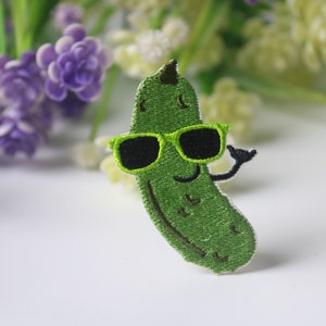 May include: Green embroidered iron-on patch of a cartoon pickle wearing sunglasses and smiling. The pickle is green and has a brown stem. The sunglasses are green with black lenses.