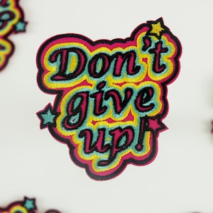 Könnte beinhalten: Ein bunter gestickter Aufnäher mit dem Text "Don't give up!" in einer Retro-Schriftart. Der Aufnäher hat ein rosa, gelbes und blaues Farbschema mit schwarzen Umrissen. Es befinden sich zwei kleine Sterne auf dem Aufnäher.