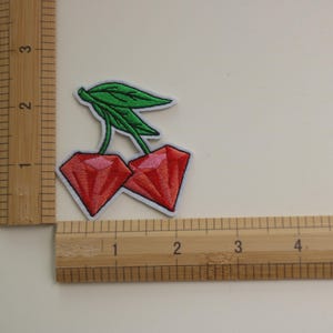 May include: Two red diamond-shaped cherries with green leaves, measured against a ruler.