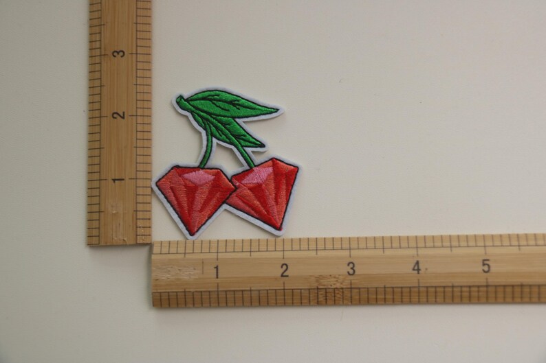May include: Two red diamond-shaped cherries with green leaves, measured against a ruler.