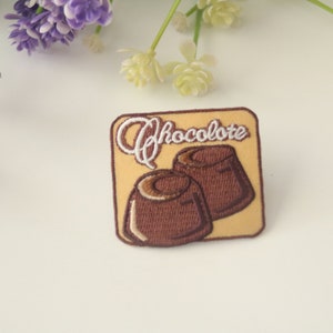 May include: A brown and yellow embroidered patch with the word "Chocolate" in white lettering and two chocolate candies.