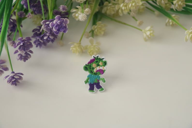 May include: Green and purple embroidered iron-on patch of a cartoon zombie with a toothy grin.