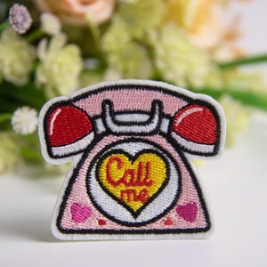 May include: Embroidered patch in the shape of a pink retro telephone with red ear pieces. The phone has a yellow heart in the center with the words "Call me". Two pink hearts are on the bottom of the phone.