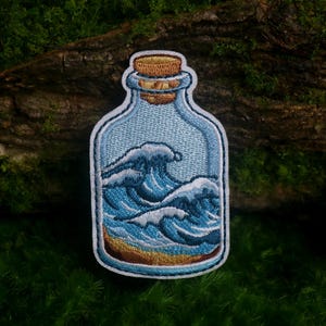 May include: An embroidered patch shaped like a glass bottle with a cork stopper. The interior of the bottle depicts a blue ocean scene with white-capped waves crashing over a sandy brown beach, all outlined in a thick white border.