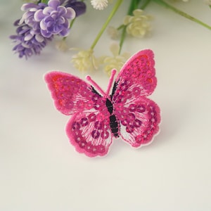 May include: A pink butterfly patch with sequin details and a black body. The butterfly is embroidered with a white border. Purple and white flowers are in the background.