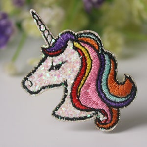 Pode incluir: Uma cabeça de unicórnio branca com glitter, com uma crina e um chifre arco-íris. O unicórnio é bordado com fio colorido.