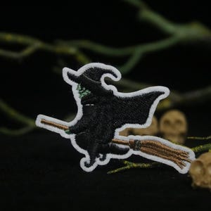 Puede incluir: Una bruja negra y verde montando una escoba sobre un fondo blanco. La bruja lleva un sombrero puntiagudo y tiene la cara verde.