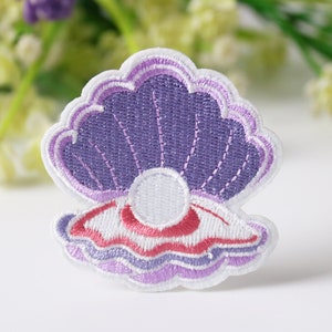 Könnte beinhalten: Gestickter Aufnäher mit einem Muscheldesign und einer Perle. Der Aufnäher hat ein lila, weißes und rotes Farbschema. Die Muschel ist geöffnet und zeigt eine weiße Perle in der Mitte. Für Kleidung oder Accessoires.