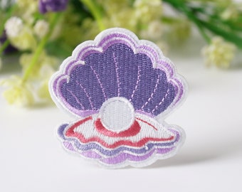 May include: Embroidered patch featuring a seashell design with a pearl. The patch has a purple, white, and red color scheme. The seashell is open, revealing a white pearl in the center. The patch is likely for clothing or accessories.
