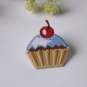 Könnte beinhalten: Ein blauer und weißer Cupcake mit braunem Frosting und einer roten Kirsche oben drauf. Der Cupcake ist auf einem weißen Stoff gestickt.