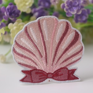 Op de afbeelding: Een roze en witte geborduurde schelp met een roze strik. De schelp is een decoratieve patch.