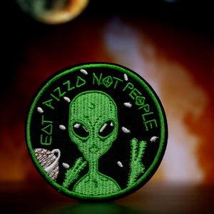 May include: A green alien with large eyes and a peace sign is on a black embroidered patch. The patch says "Eat Aliz Not People" in white text.