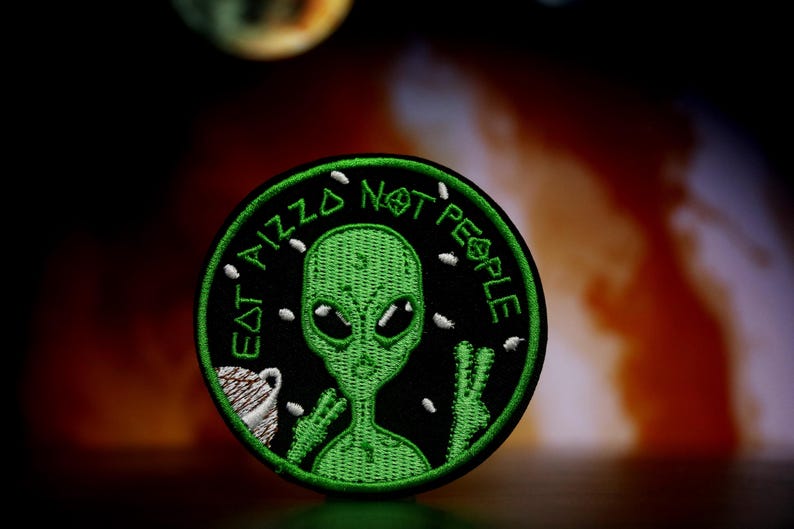 May include: A green alien with large eyes and a peace sign is on a black embroidered patch. The patch says "Eat Aliz Not People" in white text.