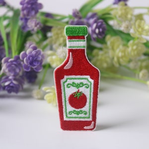 Puede incluir: Parche bordado rojo en forma de botella de ketchup con una tapa verde y una etiqueta blanca con un tomate verde.