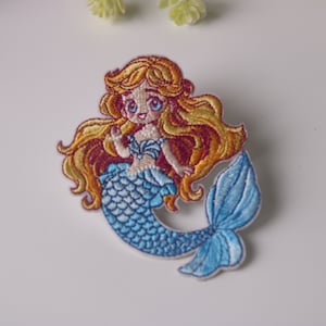 May include: Embroidered iron-on patch featuring a cartoon mermaid with long blonde hair, a blue tail, and a pink seashell bra.