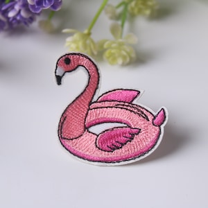 Puede incluir: Parche bordado rosa de un flamenco en un flotador inflable. El flamenco está mirando hacia la izquierda y tiene un pico rosa y patas rosas.