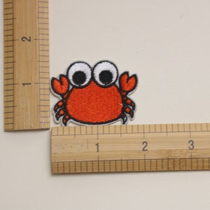 Op de afbeelding: Een geborduurde strijkpatch van een cartoonkrab met grote witte ogen. De krab is ongeveer 2,5 cm breed.