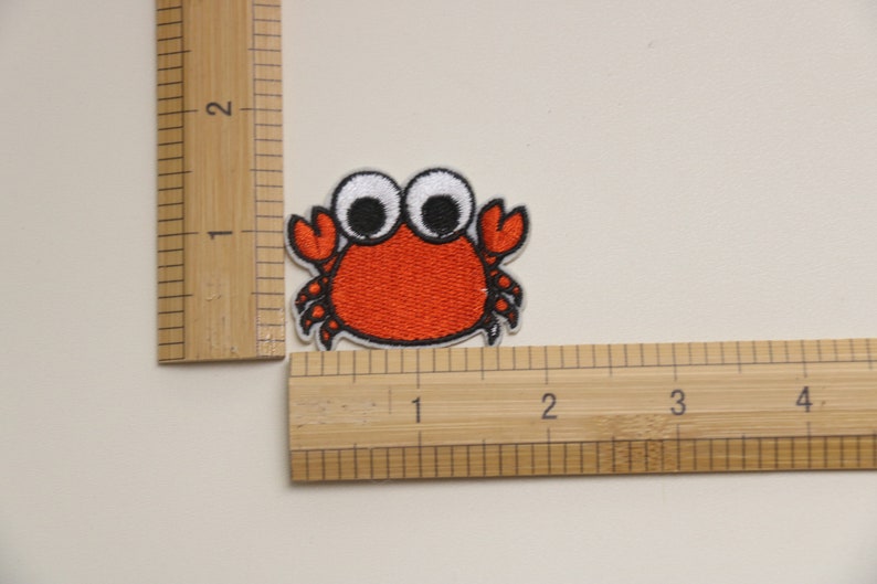 Op de afbeelding: Een geborduurde strijkpatch van een cartoonkrab met grote witte ogen. De krab is ongeveer 2,5 cm breed.