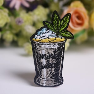 May include: Embroidered patch featuring a cocktail glass design with the words "mint juice". The patch has a black outline, a silver-colored glass, and a green leaf on top. The top of the glass has blue and yellow accents.