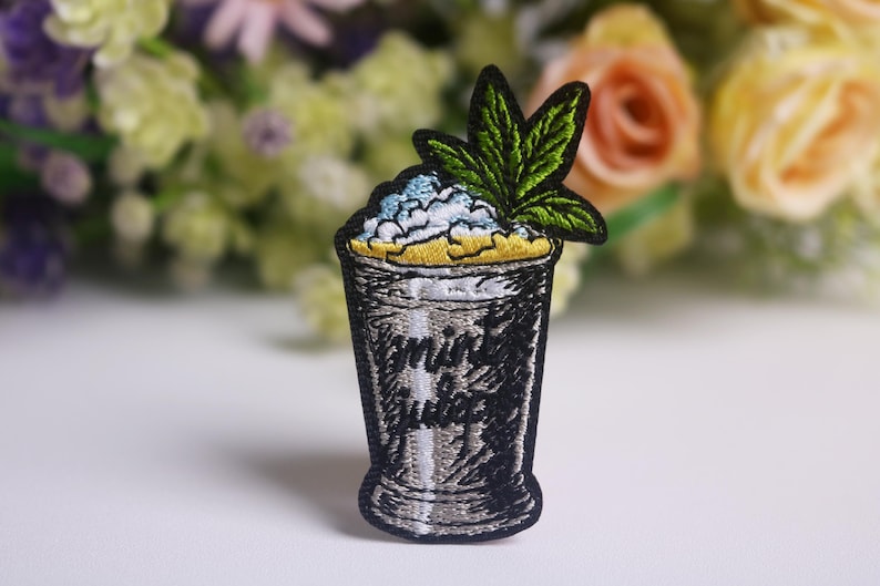 Op de afbeelding: Geborduurde patch met een cocktailglasontwerp met de tekst "mint juice". De patch heeft een zwarte omtrek, een zilverkleurig glas en een groen blad. De bovenkant van het glas heeft blauwe en gele accenten. Een decoratief item.