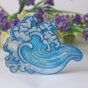 May include: A blue and white embroidered iron-on patch depicting a stylized wave. The wave is blue with white foam and a white outline.