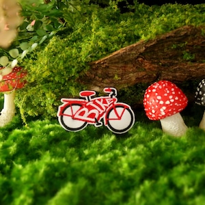 K&ouml;nnte beinhalten: Ein rotes und wei&szlig;es Spielzeugfahrrad mit dem Wort "BIKE" darauf, umgeben von gr&uuml;nem Moos und drei Spielzeugpilzen. Zwei der Pilze sind rot und wei&szlig;, und einer ist schwarz und wei&szlig;.