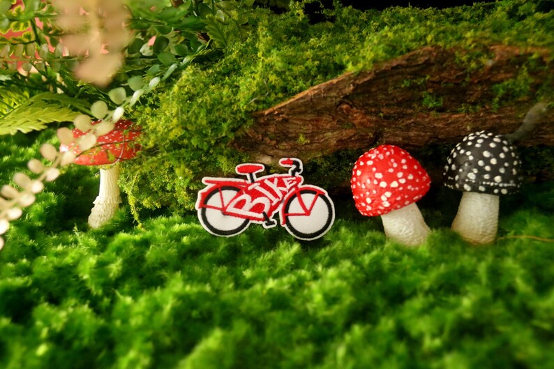 K&ouml;nnte beinhalten: Ein rotes und wei&szlig;es Spielzeugfahrrad mit dem Wort "BIKE" darauf, umgeben von gr&uuml;nem Moos und drei Spielzeugpilzen. Zwei der Pilze sind rot und wei&szlig;, und einer ist schwarz und wei&szlig;.