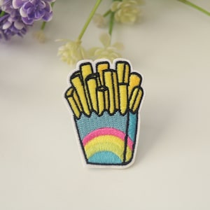 Könnte beinhalten: Gestickter Aufnäher mit einer Cartoon-Darstellung von Pommes Frites in einem blauen Behälter mit Regenbogen-Design. Die Pommes sind gelb und der Behälter hat einen weißen Rand.