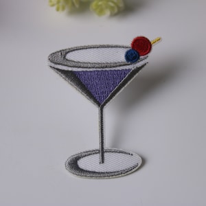May include: An embroidered iron-on patch of a purple martini glass with a red cherry and a blue blueberry on a white background.