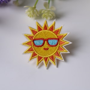 Puede incluir: Un sol amarillo bordado con una cara sonriente que lleva gafas de sol azules. El sol tiene rayos naranjas y un contorno blanco.