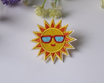 Op de afbeelding: Een geborduurde gele zon met een glimlachend gezicht dat een blauwe zonnebril draagt. De zon heeft oranje stralen en een witte omtrek.