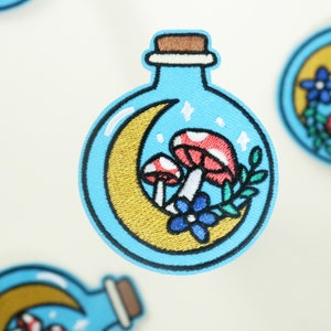 May include: A blue glass bottle with a cork stopper, containing a crescent moon, two red and white mushrooms, and a blue flower. The bottle is surrounded by a gold crescent moon.