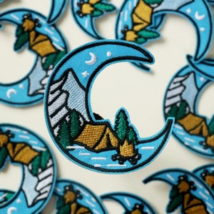 May include: A set of blue crescent moon embroidered patches with a camping scene. The scene includes a tent, mountains, trees, and a crescent moon with stars.